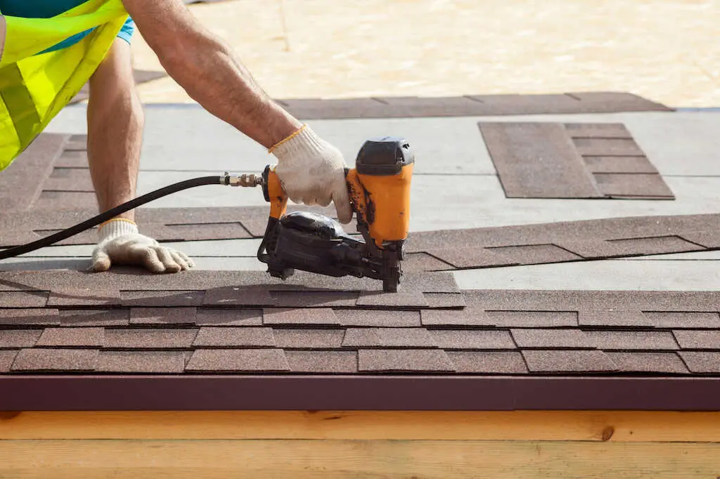 A roofer in gloves and safety gear using a nail gun for professional roofing services on a house.