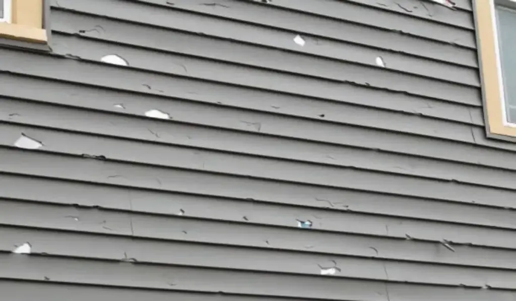 A close-up of damaged siding on a gray house, showing cracks, holes, and chipped paint from hail impact.