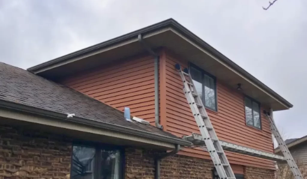 Two-story Lockport home undergoing rain gutter installation with ladders positioned along upper roofline for access.